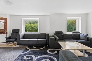 Ett modernt vardagsrum med en grå soffa, en svart fåtölj och ett soffbord. Två fönster ger utsikt över grönområden.