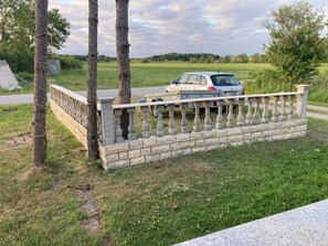 Un espace clos délimité par un muret en pierre, une voiture garée et, au fond, un champ d’herbe.