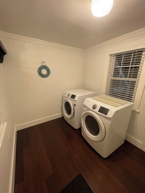 A laundry room with two front-loading washing machines, a wooden floor, a white wall with a wreath, and a window with blinds. - Quaint 2-bedroom Cottage Walking Distance to the Beach (Yarmouth)
