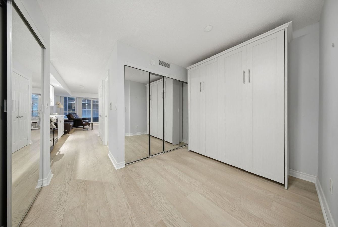 A hallway with a mirrored closet door, a wooden floor, and a view into a room with a desk and chair.