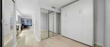 A hallway with a mirrored closet door, a wooden floor, and a view into a room with a desk and chair.