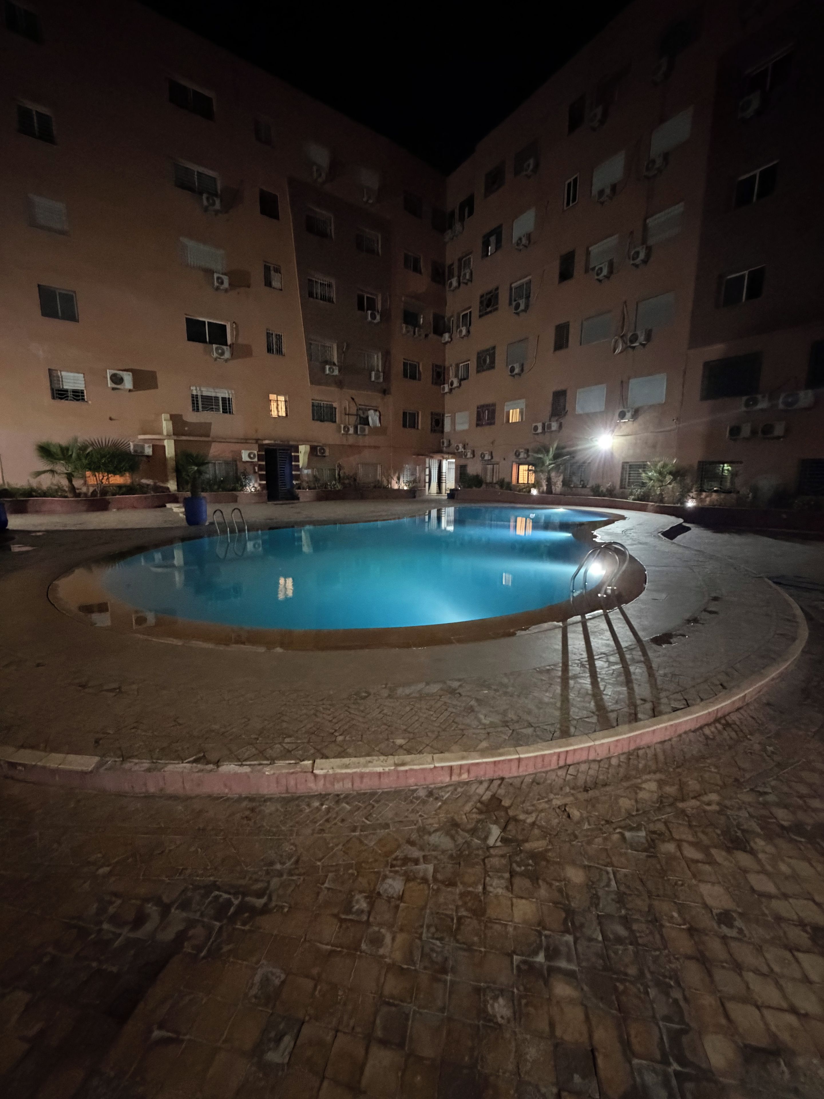 A courtyard with a swimming pool, surrounded by a brick floor and a building with multiple windows and air conditioning units.