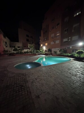A nighttime outdoor swimming pool area with a brick pathway, surrounded by buildings with multiple windows and air conditioning units. - 2bed.bathroom.2pool (Marrakech)