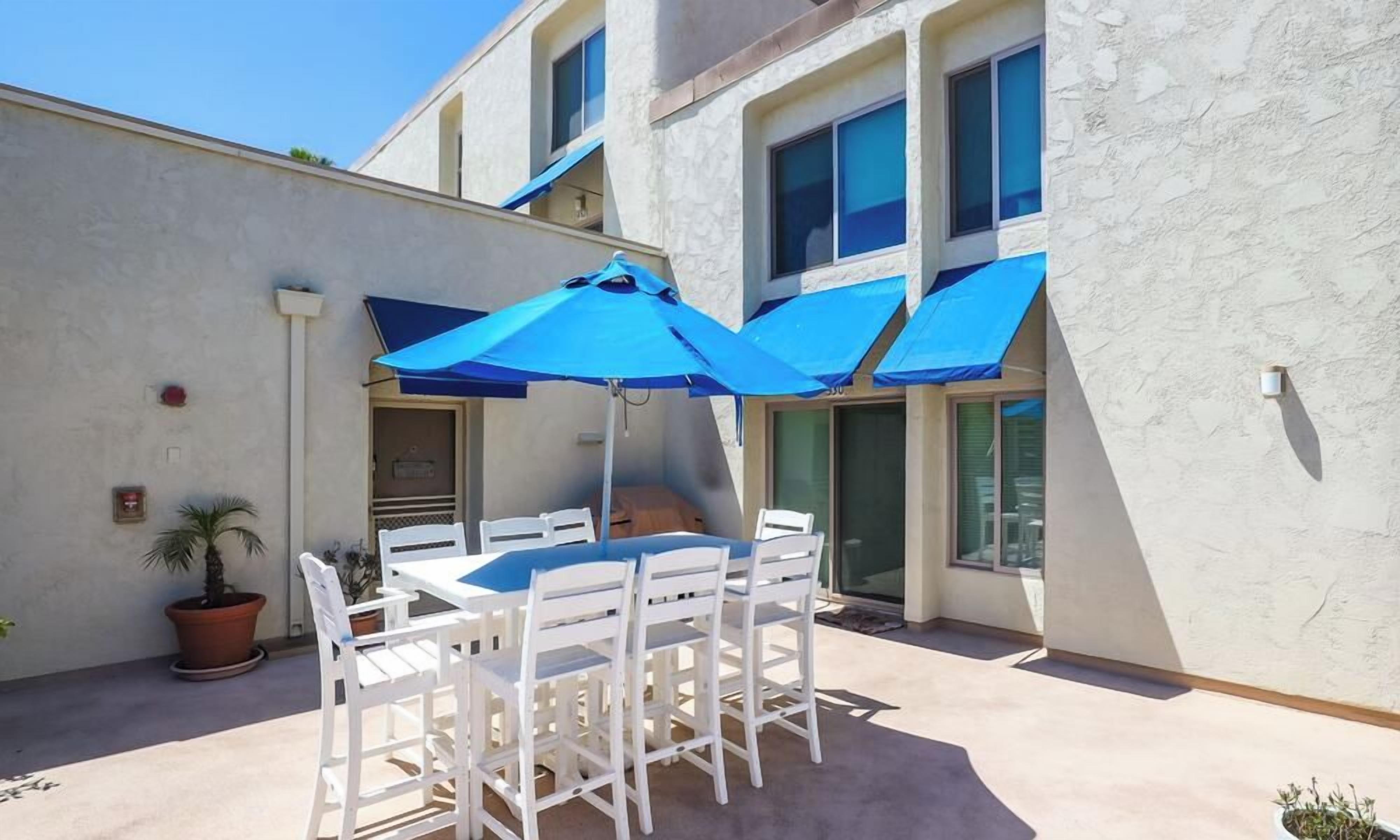 An outdoor patio with a table and chairs, blue umbrellas, and a potted plant.