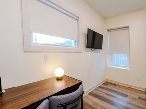 Ein Zimmer mit einem Holztisch, einer runden Tischlampe, einem an der Wand befestigten Fernseher und zwei Fenstern mit Jalousien.