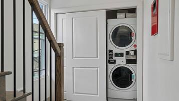 A laundry room with a front-loading washer and dryer, a wooden staircase, and a white door.