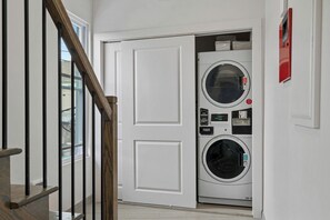 A laundry room with a front-loading washer and dryer, a wooden staircase, and a white door.