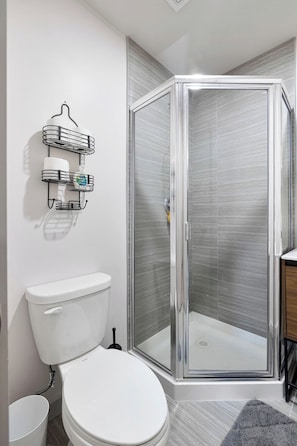 A bathroom with a white toilet, a glass-enclosed shower, and a wall-mounted shelf with toiletries. - The Columbus Hotel - The Courtyard Suite - 89-303 (Middletown)
