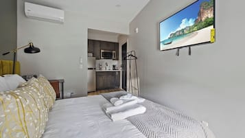 Quarto com cama, televisão, escrivaninha e uma kitchenette ao fundo.
