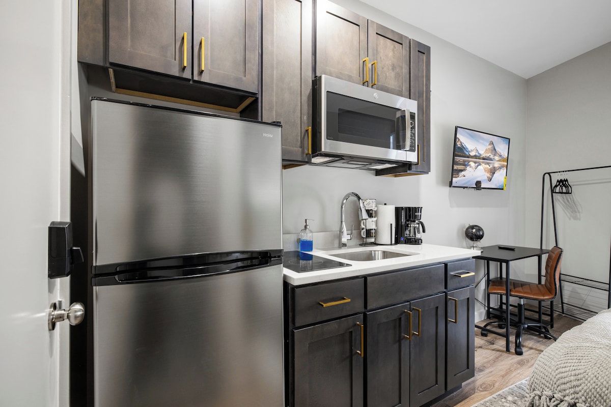 A compact kitchen with a stainless steel refrigerator, a microwave, and a coffee maker. There is a small table with a chair and a painting on the wall.