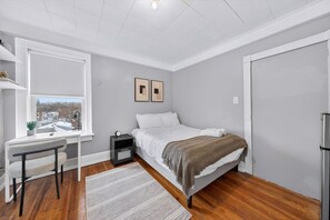 Ein Schlafzimmer mit Bett, Schreibtisch mit Stuhl, Nachttisch, Fenster und Tür.