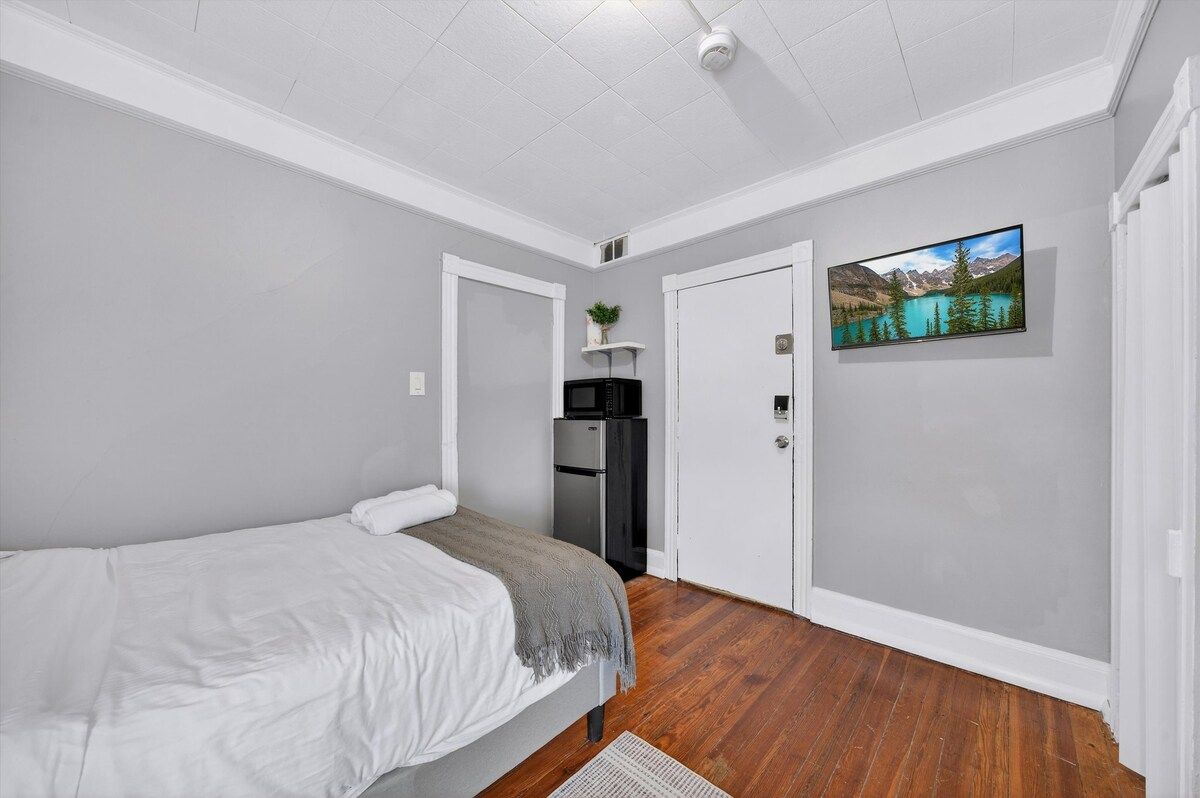 Un dormitorio con cama, refrigerador, televisor y puerta que da a otra habitación.