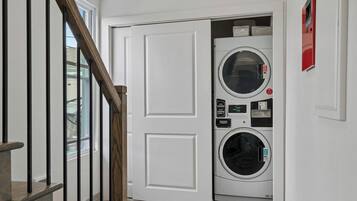 A laundry room with a front-loading washer and dryer, a wooden staircase, and a white door.