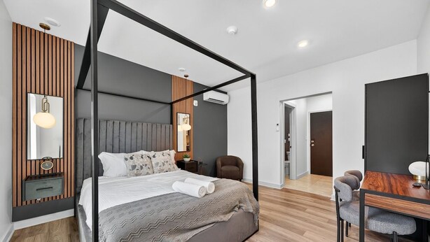 A modern bedroom with a four-poster bed, a wooden accent wall, a bedside table with a lamp, and a view into the adjacent dining area. - The Columbus Hotel - The Canopy Studio - 52-301 (Middletown)