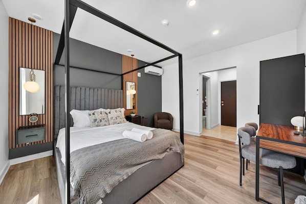 A modern bedroom with a four-poster bed, a wooden accent wall, a bedside table with a lamp, and a view into the adjacent dining area. - The Columbus Hotel - The Canopy Studio - 52-301 (Middletown)