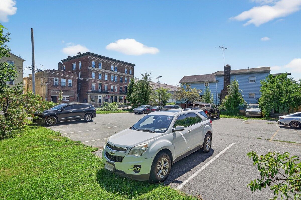 A parking lot with several cars, a grassy area, and buildings in the background.