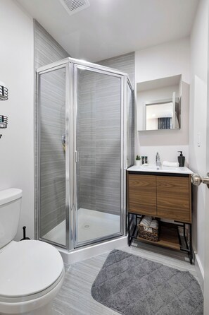 A modern bathroom with a glass shower enclosure, a white toilet, a wooden vanity, and a mirror above the sink. - The Columbus Hotel - The Scholar Suite - 89-307 (Middletown)