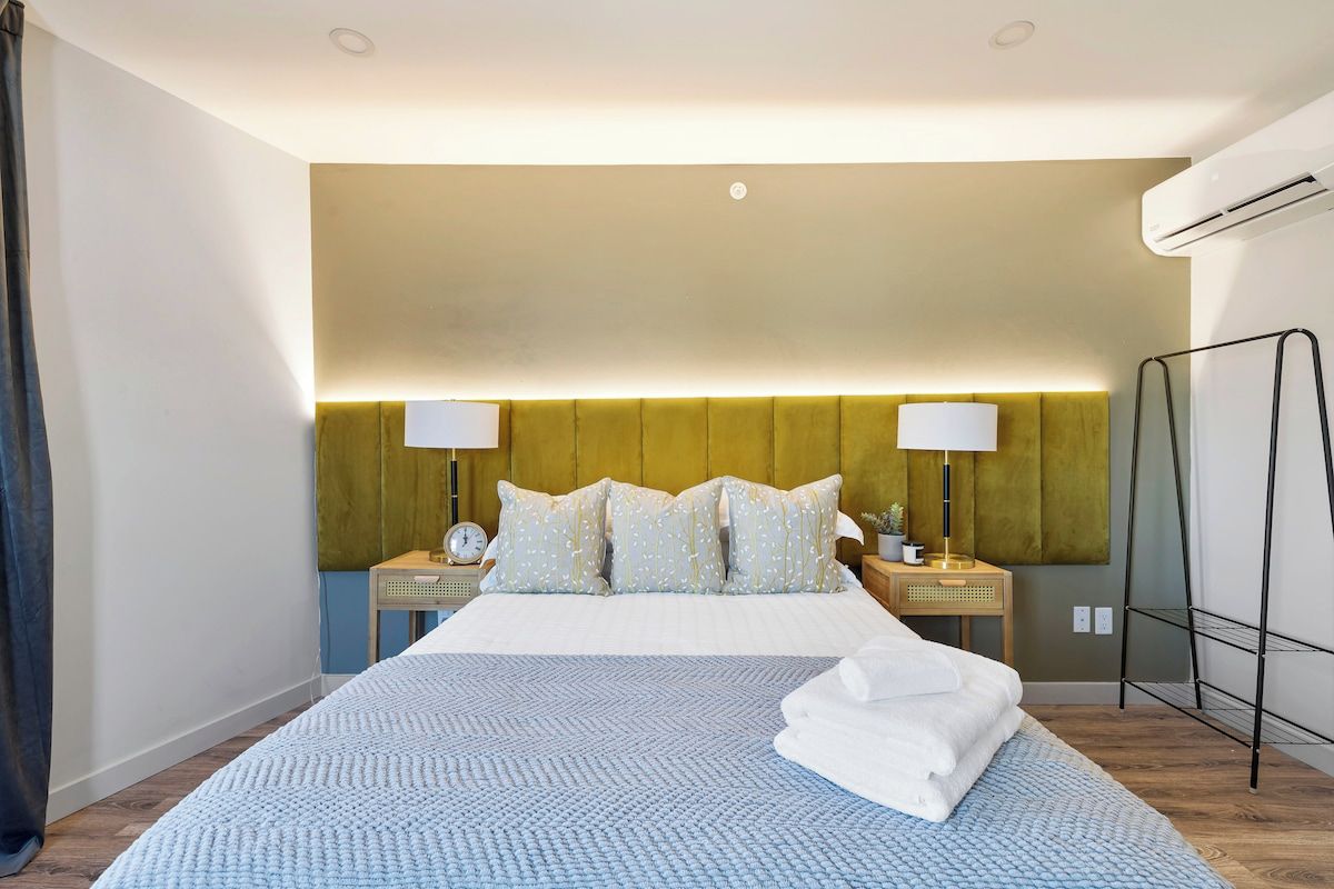 A hotel room with a large bed, two bedside tables, a wall-mounted air conditioning unit, and a floor lamp.