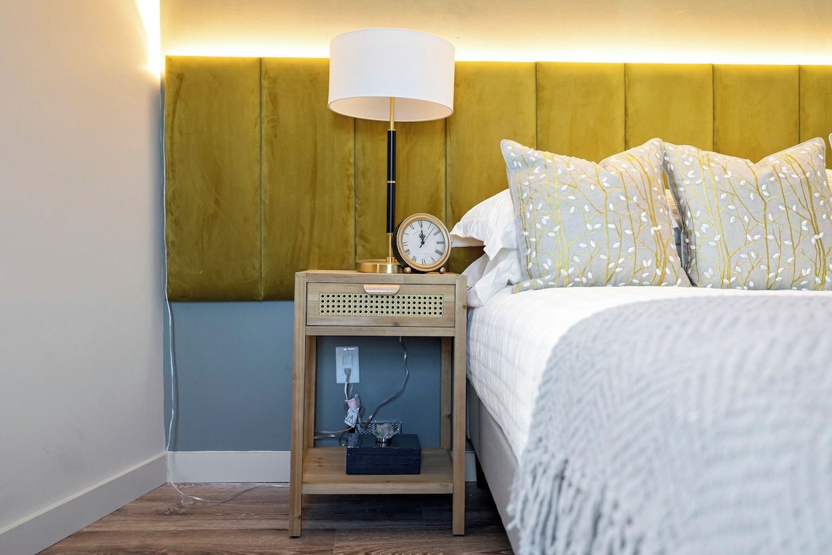 A neatly made bed with patterned pillows, a bedside lamp, and a small wooden nightstand with a clock and a power outlet.