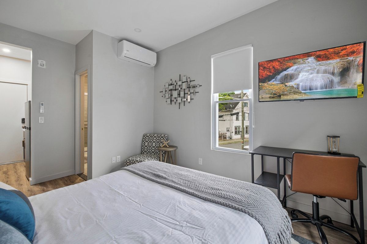 Een slaapkamer met een bed, bureau, stoel, raam met uitzicht en een tv aan de muur.