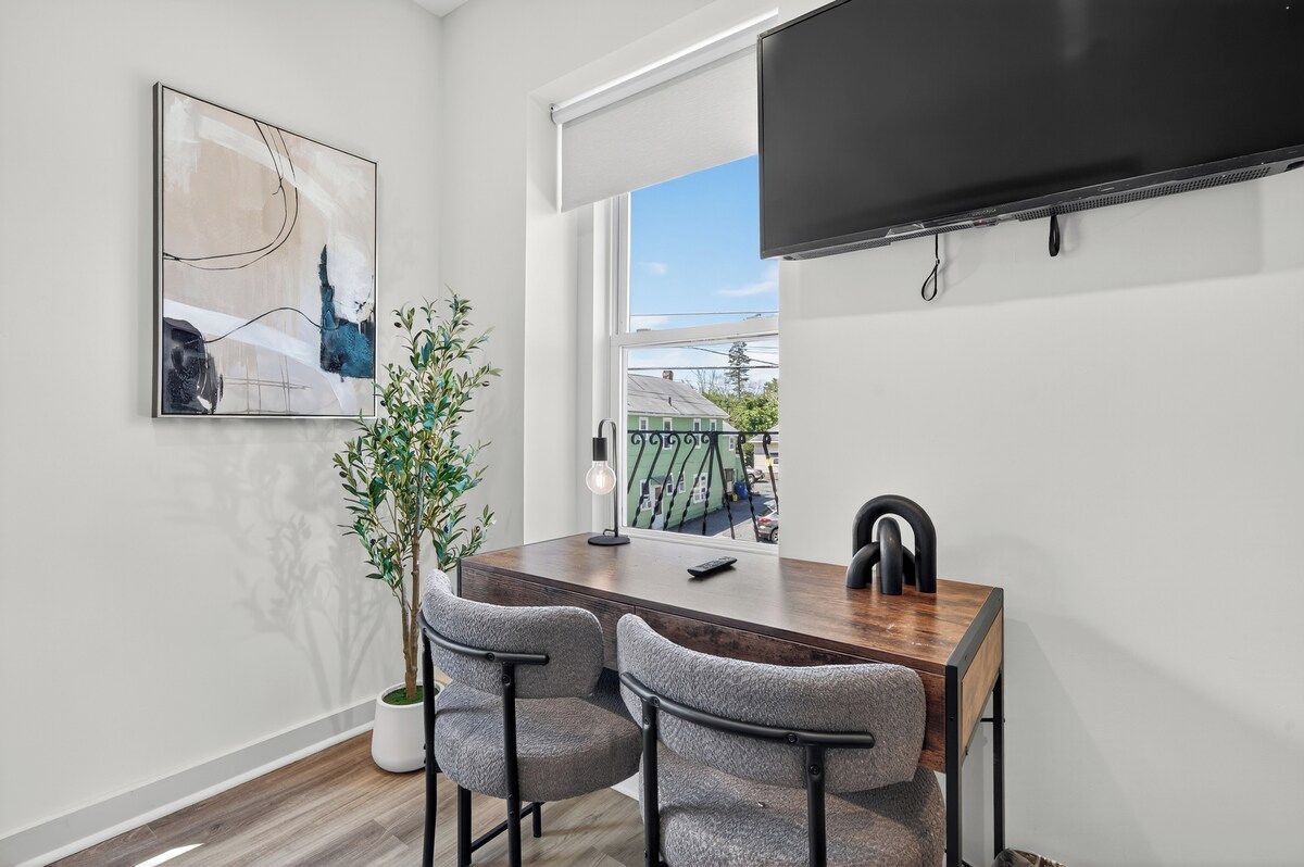 A modern dining area with a wooden table, two grey chairs, a potted plant, and a flat-screen TV mounted on the wall.