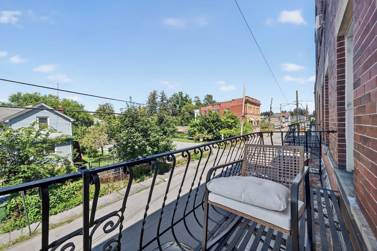 A balcony with a metal railing, a single cushioned bench, and a view of a street with houses and trees.