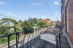 A balcony with a metal railing, a single cushioned bench, and a view of a street with houses and trees.