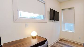 A room with a wooden desk, a round table lamp, a mounted TV, and two windows with blinds.