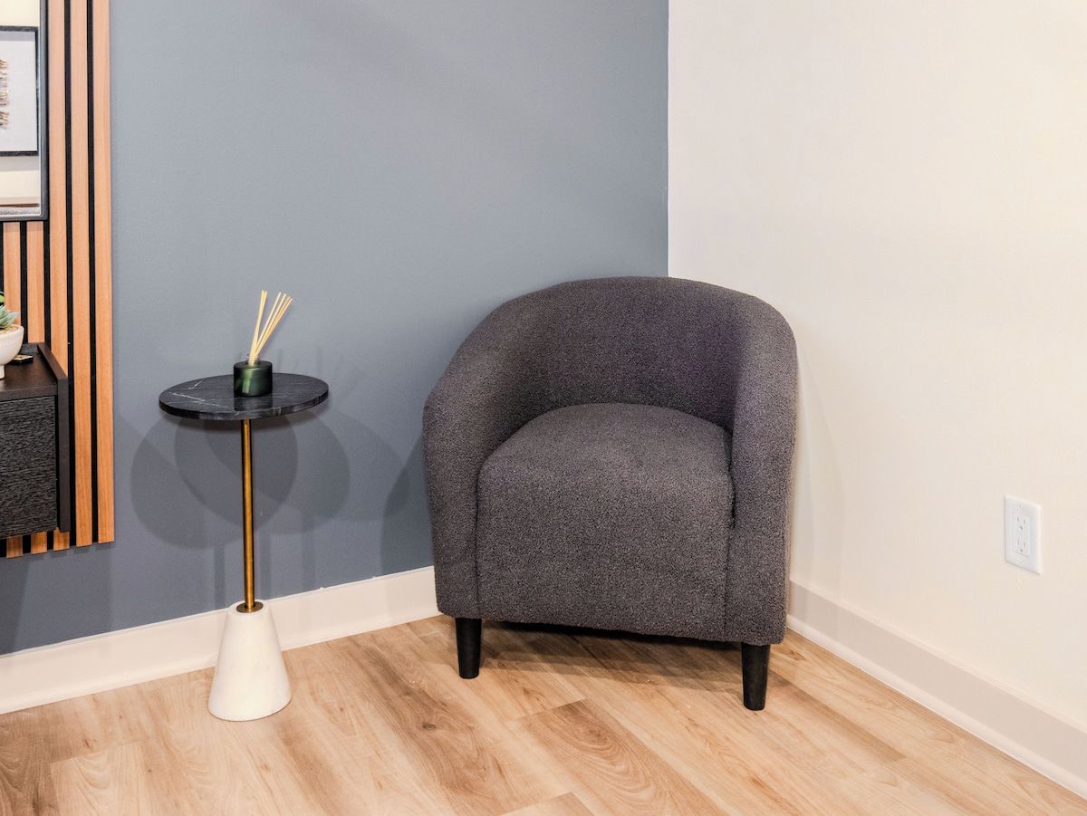 A room with a grey armchair, a small round table with a black top and gold legs, and a white conical base.