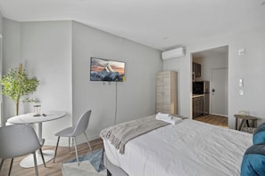 Ett modernt hotellrum med en säng, en tv som är monterad på väggen, ett litet matbord med stolar, en krukväxt och utsikt över köket.