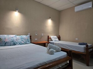 Quarto com duas camas de solteiro, cada uma com estrutura de madeira e roupa de cama branca. Há luminárias fixadas na parede acima de cada cama, e um ar-condicionado na parede.