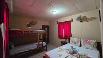 A dormitory room with bunk beds, a single bed, a fan, a clock, and a window with red curtains.