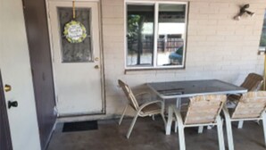Un patio con una mesa y sillas, una puerta decorada con un adorno colgante, y una ventana que ofrece vistas al exterior.