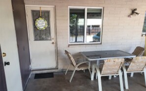 Une terrasse avec une table et des chaises, une porte ornée d’une décoration suspendue et une fenêtre donnant sur l’extérieur.