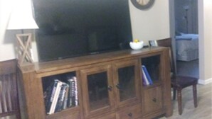Una sala de estar con un televisor sobre un mueble de madera, un reloj en la pared y un acceso a otra habitación.