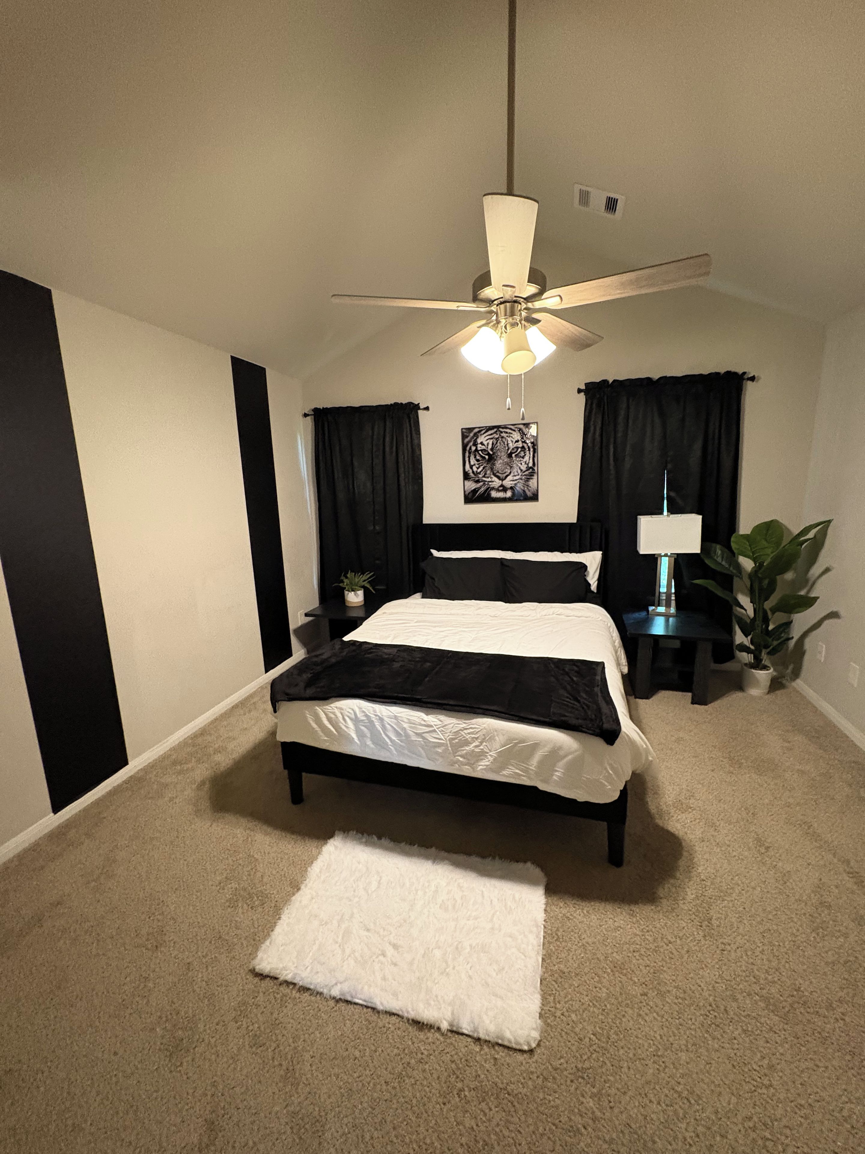 A bedroom with a bed, a ceiling fan, a nightstand, a lamp, and a potted plant.