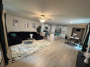 Een moderne woonkamer met een zwarte modulaire bank, een vloerkleed met zebraprint en een keukenhoek met witte kastjes en een barkruk.