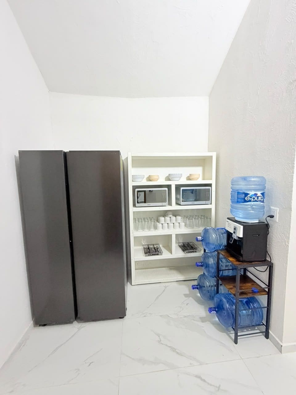A compact kitchen with a double-door refrigerator, a white shelving unit with kitchenware, and a water dispenser.