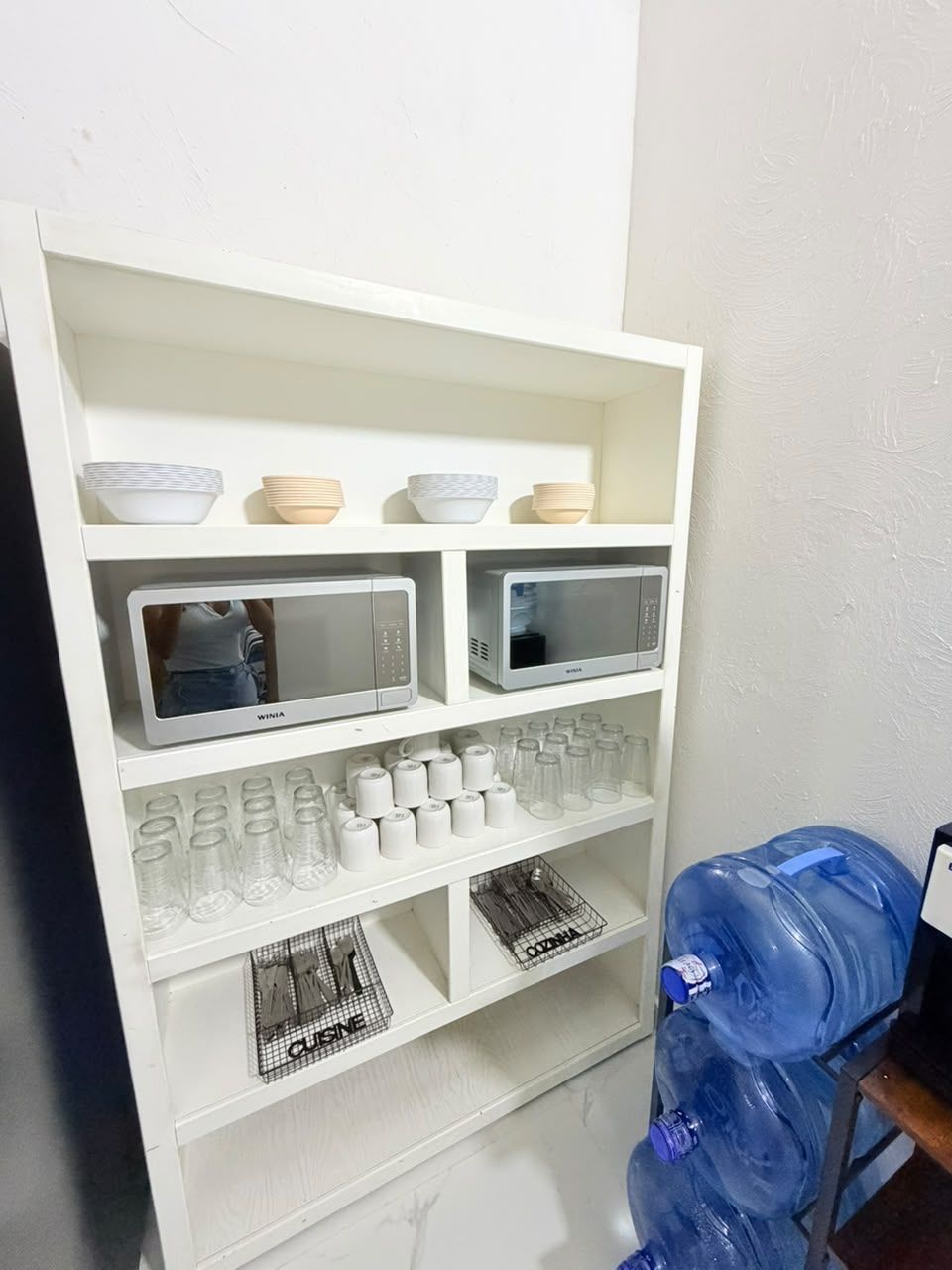 Un mueble blanco con microondas, cristalería y dispensador de agua.
