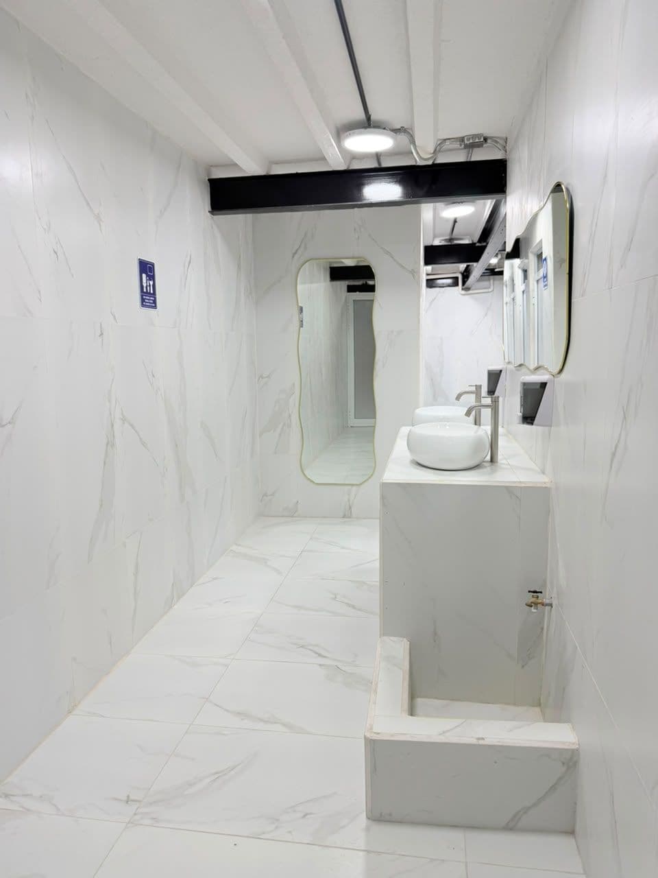 A modern bathroom with a white marble floor and walls, a white sink with a mirror above it, and a shower area visible in the background.