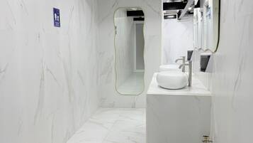 A modern bathroom with a white marble floor and walls, a white sink with a mirror above it, and a shower area visible in the background.