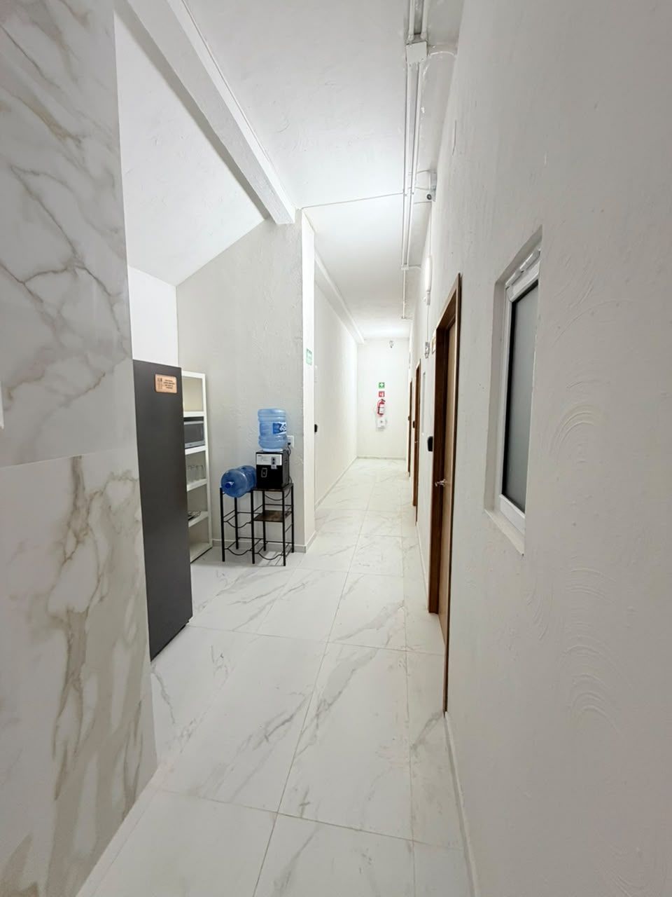 Un pasillo con paredes blancas y suelo de mármol, que conduce a una habitación con cama y ventana.