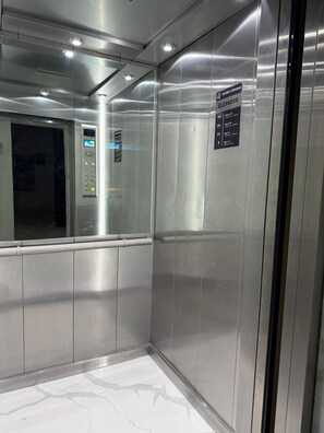 Interior de un ascensor con paredes metálicas, un panel digital y un letrero que indica la capacidad máxima y los niveles de los pisos.