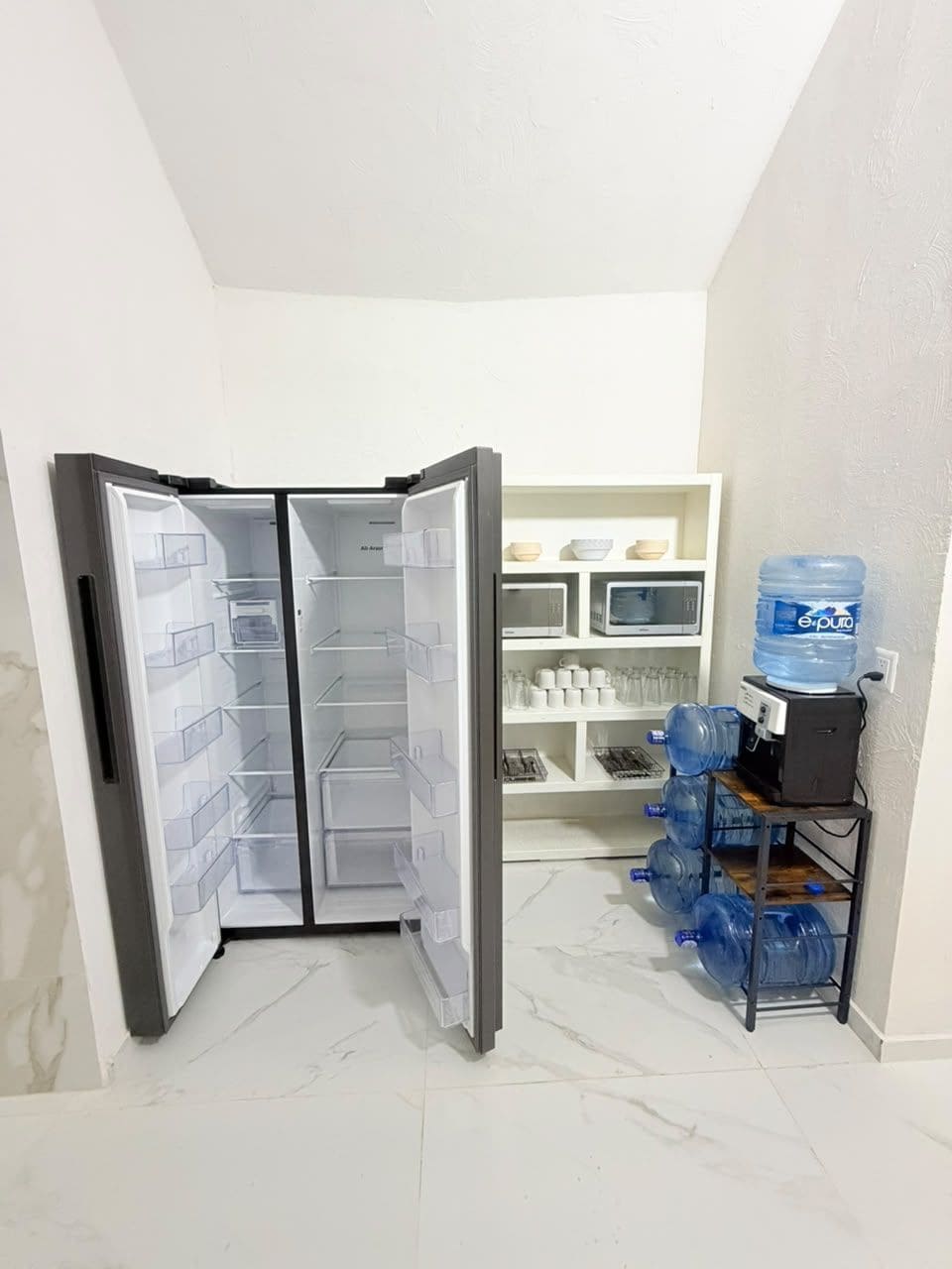 A kitchen with a refrigerator, water dispenser, and shelves with kitchenware.
