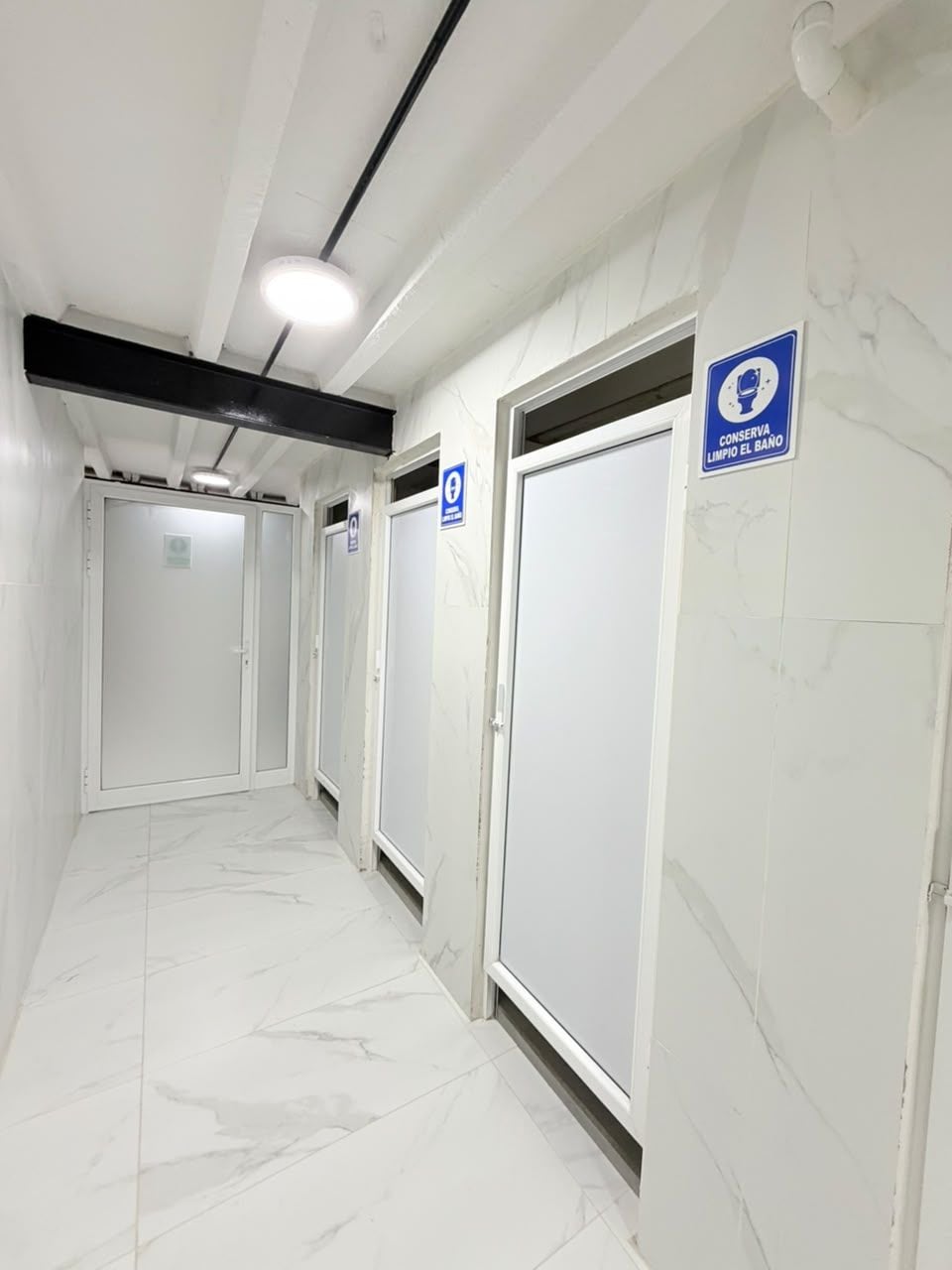 A corridor with white tiled floors and doors, some marked with blue signs indicating facilities.