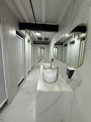 A long, narrow bathroom with white marble floors and walls, a central sink with a large mirror, and a black soap dispenser. - Palace Orange (Guadalajara)