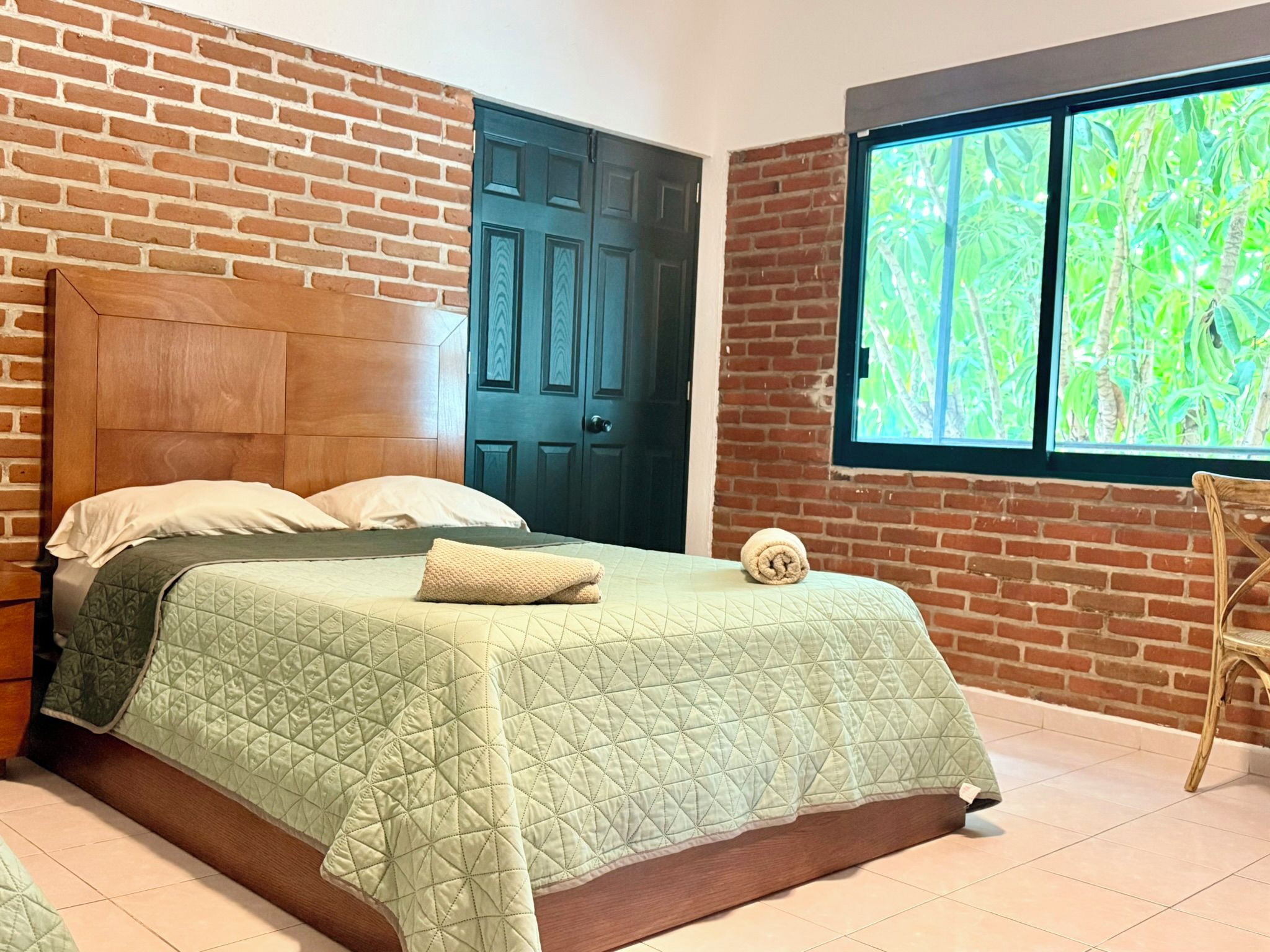 A bedroom with a brick wall, a wooden headboard, a bed with green bedding, a window with a view of greenery, and a door.
