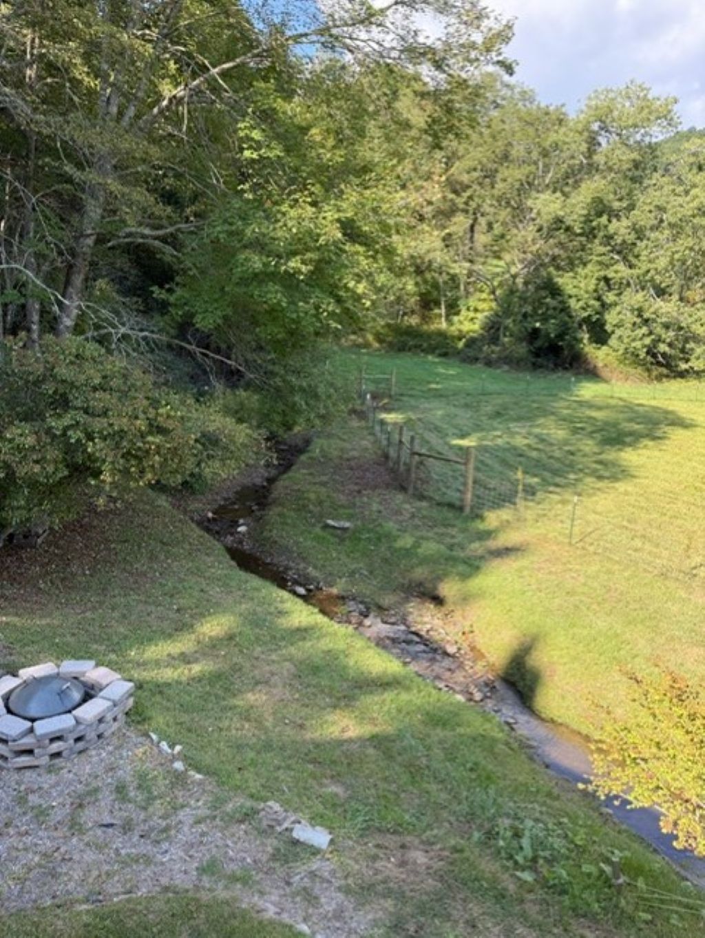 A grassy area with a small stream, a wooden fence, and a stone fire pit.