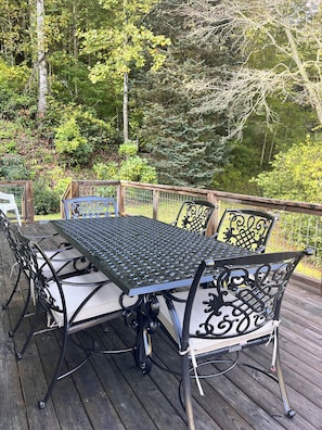 A deck with a metal table and chairs, surrounded by a wooden railing and greenery. - MOUNTAINSTEAD 3BD Glenville-Cashiers Farmhouse Big views creek waterfall donkeys (Glenville)