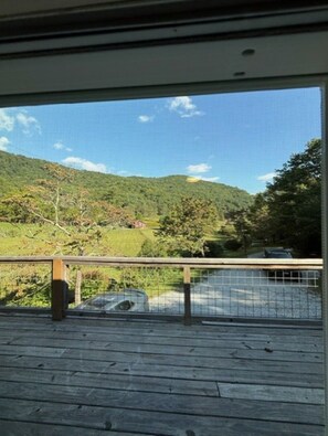A view from a window with a wooden deck, overlooking a green landscape with hills and a clear sky. - MOUNTAINSTEAD 3BD Glenville-Cashiers Farmhouse Big views creek waterfall donkeys (Glenville)